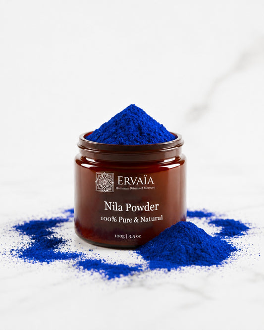 A jar of Ervaia Nila Powder skincare product to brighten and reduce the hyperpigmentation on face and body with vibrant blue powder piled on top and scattered around it on a white marble surface. The product is in a dark amber glass jar with a brown label, and the brand name 'ERVAia' is clearly visible.